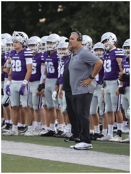 A group of people in purple and white football uniforms stand in a line on a field next to a person wearing a gray shirt, black pants, and a headset. Description generated by AI
