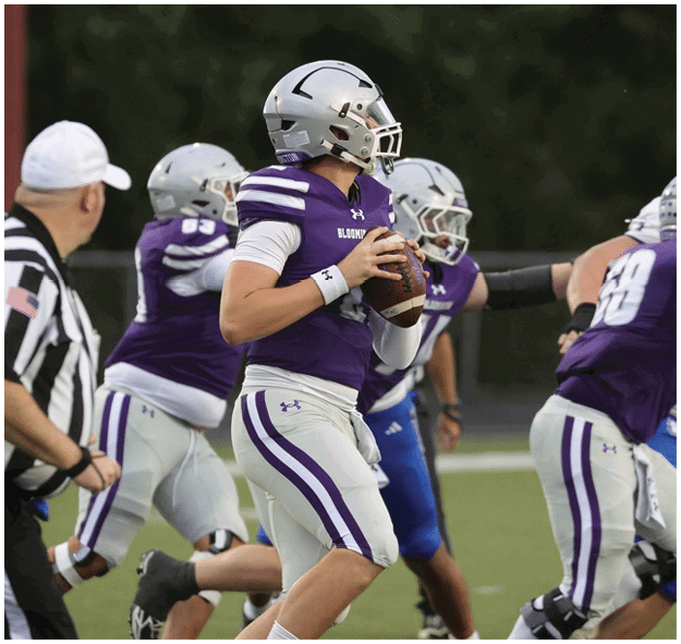 A person in a purple and white football uniform is holding a football and preparing to throw while a referee and other players are nearby on a football field. Description generated by AI