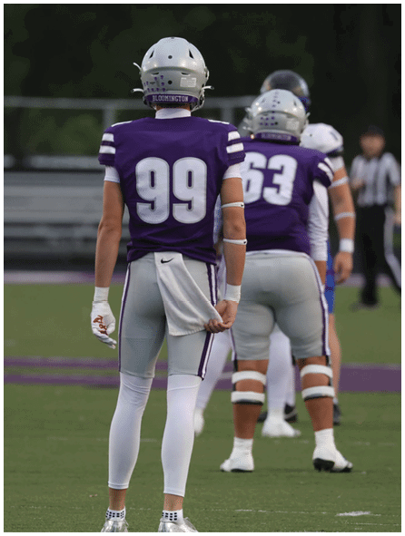 A person wearing a purple football jersey with the number 99 and a helmet labeled Bloomington stands with another person in a similar uniform in the background. Description generated by AI