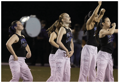 Several people wearing black sleeveless tops and light-colored pants are performing a coordinated dance or cheer routine on a field. Description generated by AI