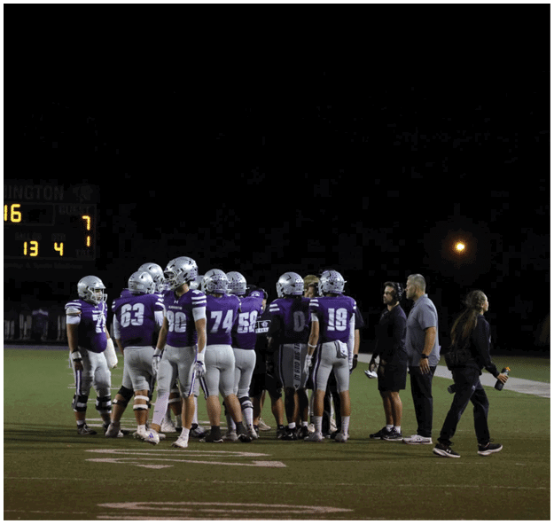 A group of football players in purple and white uniforms and coaches are gathered on a field at night with a scoreboard visible in the background. Description generated by AI