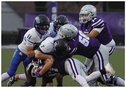 Several people wearing football uniforms are engaged in a tackle during a game on a grassy field. Description generated by AI