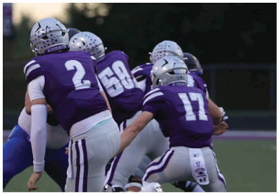 People wearing purple and white football uniforms are engaged in a play on a football field. Description generated by AI
