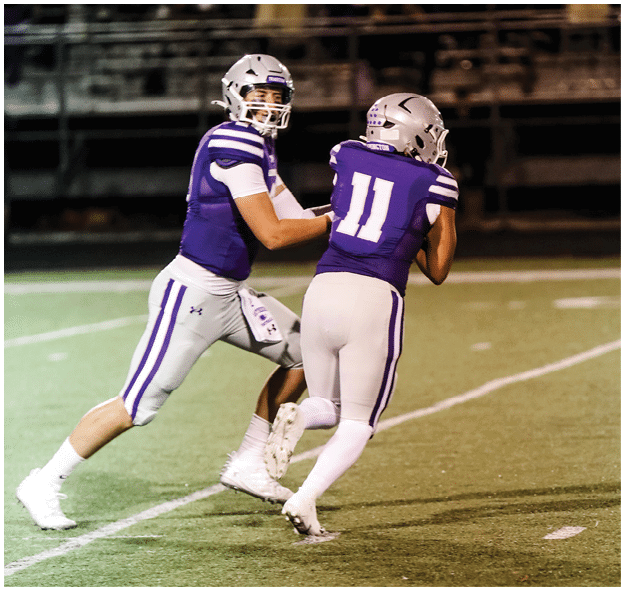 Two people wearing purple and white football uniforms are on a field, with one person handing off the ball to the other. Description generated by AI