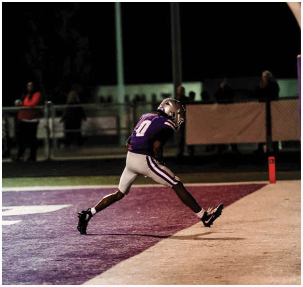 A person in a purple sports uniform and helmet is running on a field at night with spectators in the background. Description generated by AI