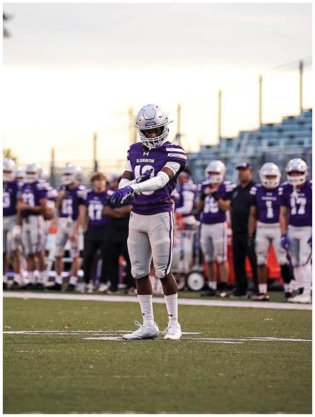 A person in a purple and white football uniform stands on a field with a group of similarly dressed people and empty bleachers in the background during sunset. Description generated by AI