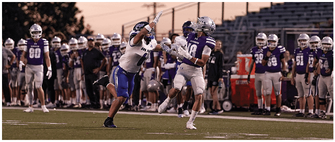 Two people in football uniforms are actively reaching for a football during a game with a crowd of players and coaches in the background under a dusky sky. Description generated by AI