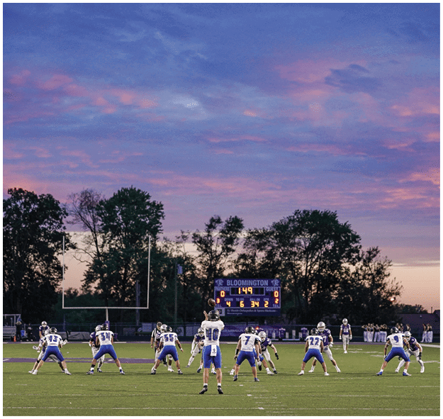 A group of people in sports uniforms are positioned on a football field during a game with a scoreboard and trees visible in the background at sunset. Description generated by AI