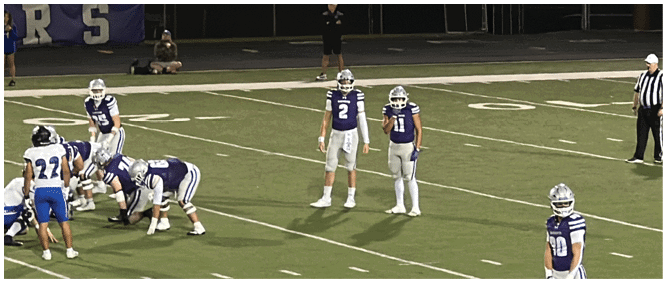 People in football uniforms are positioned on a field preparing for a play during a game. Description generated by AI