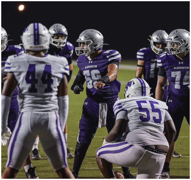 Several people wearing purple and white football uniforms and helmets are gathered on a field under a dark sky. Description generated by AI