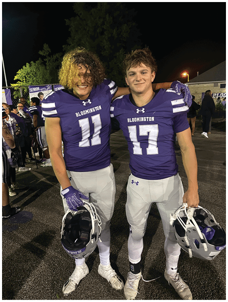 Two people wearing purple Bloomington football jerseys with numbers 11 and 17 are smiling with their arms around each other at night. Description generated by AI