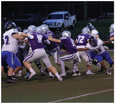 Several people wearing football helmets and uniforms are engaged in a play on a football field at night with a white truck visible in the background. Description generated by AI