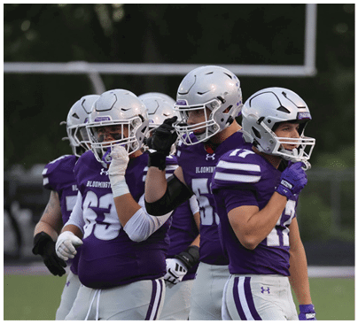 Several people wearing white football helmets and purple jerseys are gathered closely, with one person adjusting their helmet. Description generated by AI