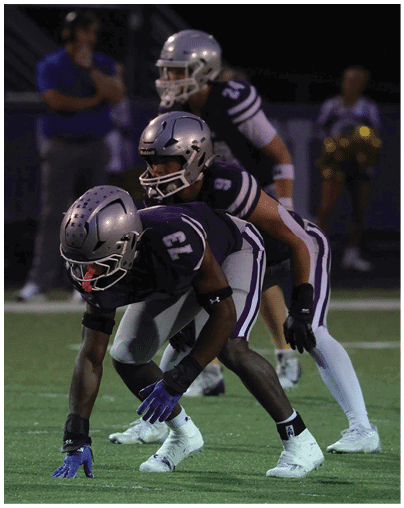 Three people wearing silver helmets and purple sports uniforms are positioned closely together, likely preparing for a play in a nighttime football game. Description generated by AI