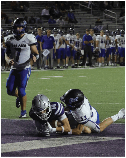 Two people in football uniforms are on the ground near the end zone while another person in a football uniform runs toward them on a field with spectators in the background. Description generated by AI