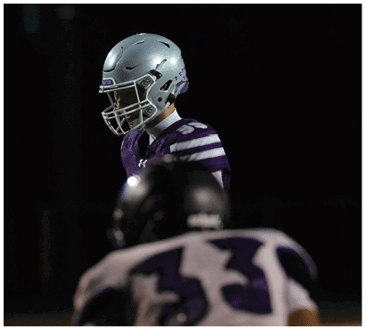 A person wearing a silver football helmet with a faceguard is shown against a dark background. Description generated by AI