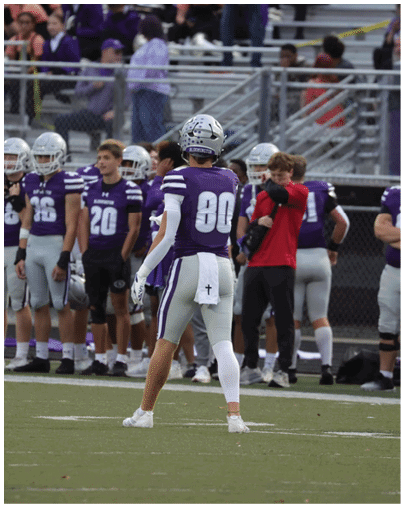 Two people in football uniforms are on the ground near the end zone while another person in a football uniform runs toward them on a field with spectators in the background. Description generated by AI