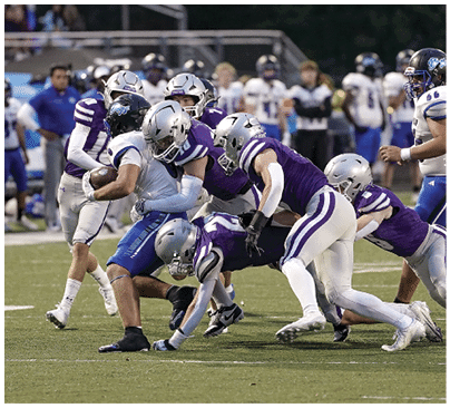A group of people in football uniforms are engaged in a tackle on a grassy field during a game. Description generated by AI