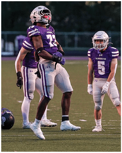 Three people in purple and white football uniforms stand on a field, with one person in mid-jump and the others watching. Description generated by AI