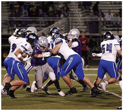 A group of people wearing football uniforms are actively engaged in a play on a football field with some players blocking and others attempting to tackle. Description generated by AI