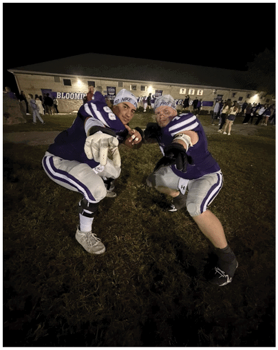 Two people wearing purple and white football uniforms and headbands are crouching and pointing towards the camera on a grassy field at night with a building and crowd in the background. Description generated by AI