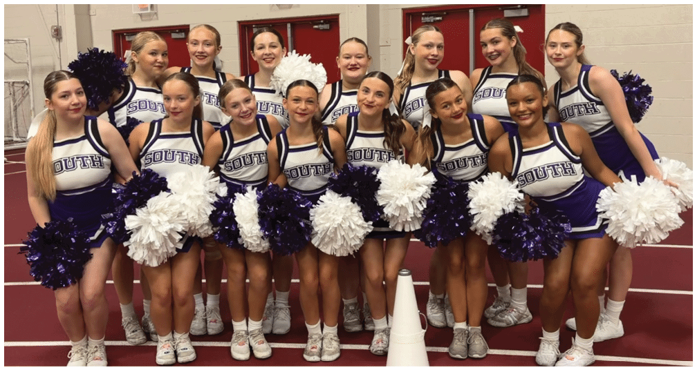 A group of people dressed in matching cheerleading uniforms holding white and purple pom-poms are standing closely together indoors. Description generated by AI