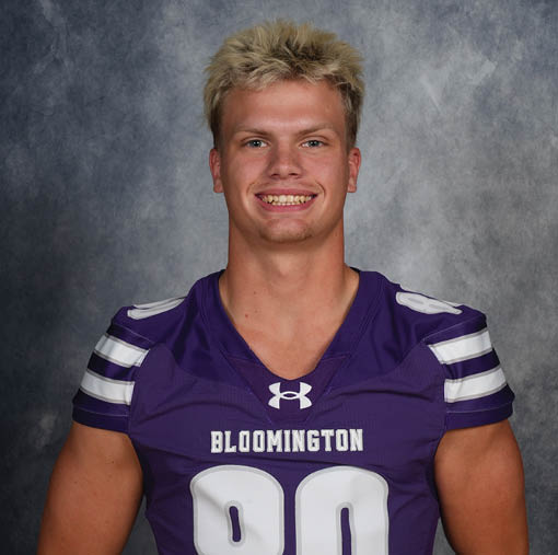 Person with short blond hair wearing a purple Bloomington sports jersey smiling against a gray textured background. Description generated by AI