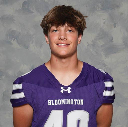 A person with short tousled hair is smiling and wearing a purple sports jersey with the word Bloomington and the number 40 on it against a gray textured background. Description generated by AI
