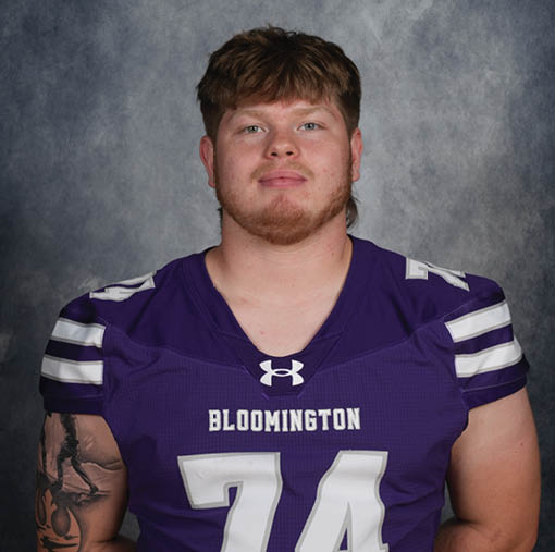 A person with short, tousled hair wearing a purple Bloomington sports jersey with the number 74 and a tattoo on their left arm is posing against a gray mottled background. Description generated by AI