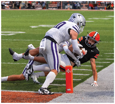 A person in a black and orange football uniform is reaching toward the end zone while two people in white and purple uniforms attempt to tackle them on a football field. Description generated by AI