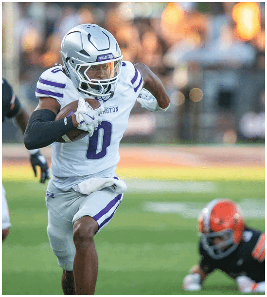 A person wearing a white and purple football uniform is running with the ball while another person in a black and orange uniform chases behind on a grassy field. Description generated by AI