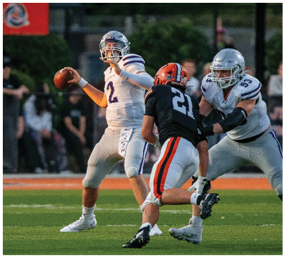 A person in a white football uniform is preparing to throw a football while being approached by a person in a black and orange uniform and another person in a white uniform on a grassy field. Description generated by AI
