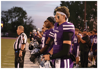 People in football uniforms and a referee stand attentively on the sidelines of a football field during a game at dusk. Description generated by AI