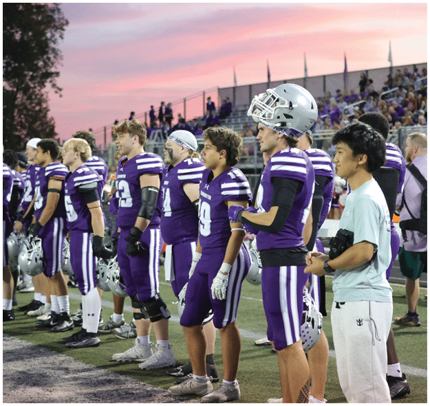 A group of people wearing purple and white football uniforms and one person in casual clothing stand in a line on a sports field during sunset with spectators in the background. Description generated by AI