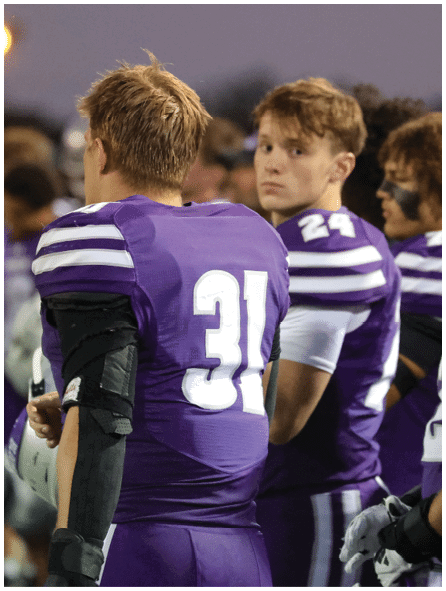 Two people wearing purple football uniforms with white numbers stand on a field, one facing away and the other looking toward the camera. Description generated by AI
