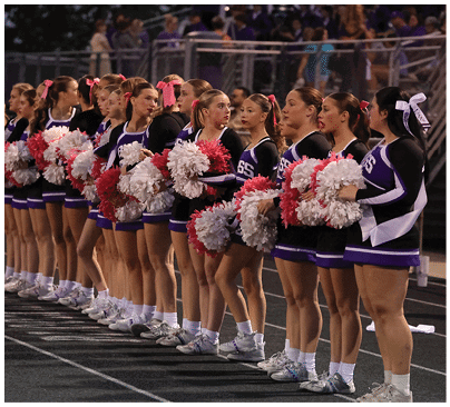A line of cheerleaders in matching uniforms with pink hair bows stand on a track holding red and white pom-poms during an outdoor event. Description generated by AI