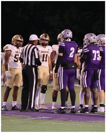 A group of football players in purple and white uniforms and a referee are standing together on a field during a game. Description generated by AI