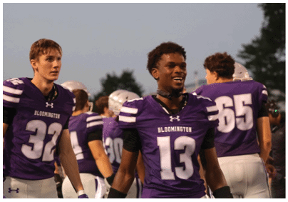 Adults wearing purple and white football uniforms with “Bloomington" on the jerseys are standing on a field during an outdoor event at dusk. Description generated by AI