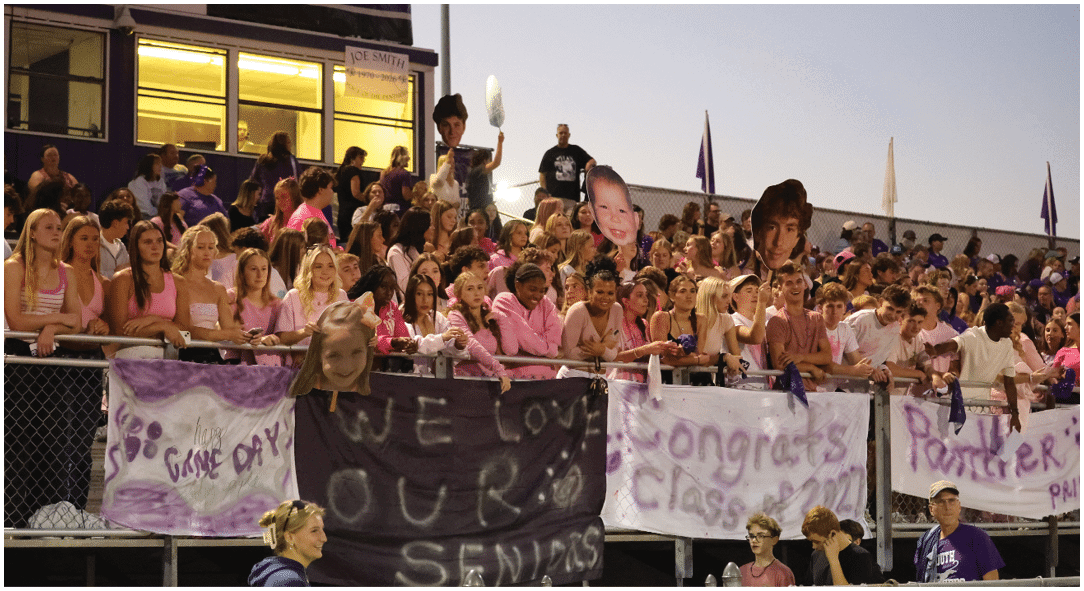 A crowd of people in bleachers at an outdoor event hold large cutout faces and banners with supportive messages for seniors. Description generated by AI