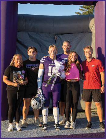 Three people stand smiling next to a large purple inflatable structure with the word “PANTHERS" on it, with one person holding a football helmet and another holding flowers. Description generated by AI