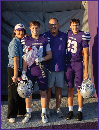 Four people stand closely together smiling with two wearing purple football uniforms holding helmets and flowers, and two wearing casual clothing, against a dark backdrop. Description generated by AI
