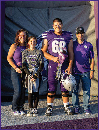Four people stand together smiling on a sports field with one person wearing a football uniform holding a helmet and flowers while the others wear casual clothing and hold flowers. Description generated by AI