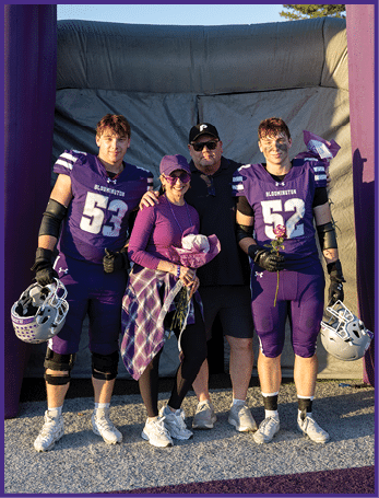 Two people in purple football uniforms holding helmets stand on either side of two people dressed in casual clothing, all smiling and posing together outdoors near an inflatable structure. Description generated by AI