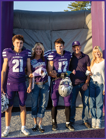 Two people in purple football uniforms stand with three casually dressed adults smiling outdoors in front of an inflatable backdrop. Description generated by AI