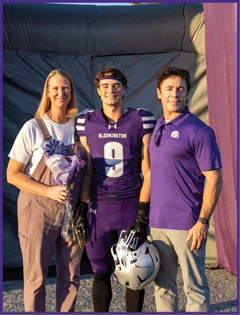 Three people stand together smiling with one person in a purple football uniform holding a helmet and the other two wearing casual clothing, one holding a bouquet of flowers, against a backdrop. Description generated by AI