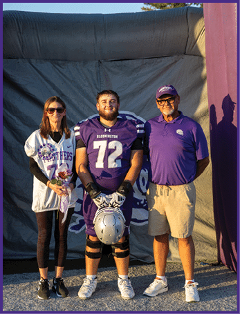 Three people stand side by side outdoors with one person in a football uniform holding a helmet, another person holding flowers, and the third person wearing a purple shirt and cap. Description generated by AI