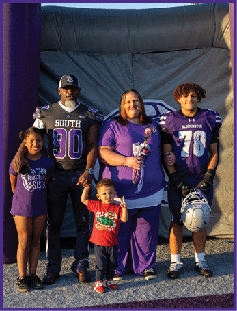 Four people stand together outdoors with two wearing football uniforms, one holding flowers, and a child making a peace sign. Description generated by AI