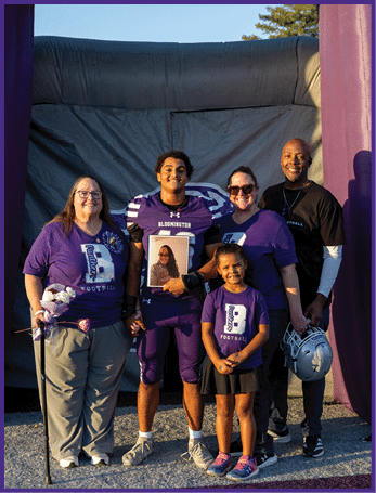 A group of five people stand together smiling outdoors with one person in a football uniform holding a framed photo while others wear matching purple shirts and one holds a bouquet of flowers. Description generated by AI