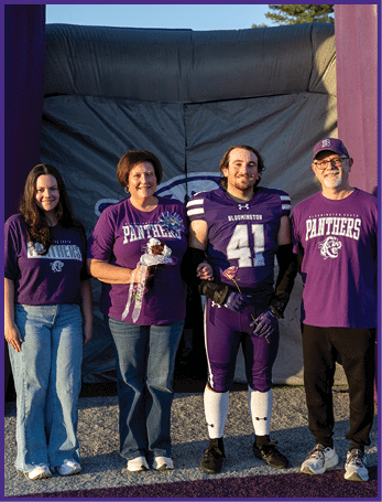 Three people stand side by side outdoors with two wearing purple shirts and one in a purple football uniform holding a flower. Description generated by AI