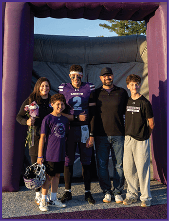 Five people stand together smiling under a large purple inflatable arch, with one person wearing a football uniform and another holding a bouquet of flowers. Description generated by AI